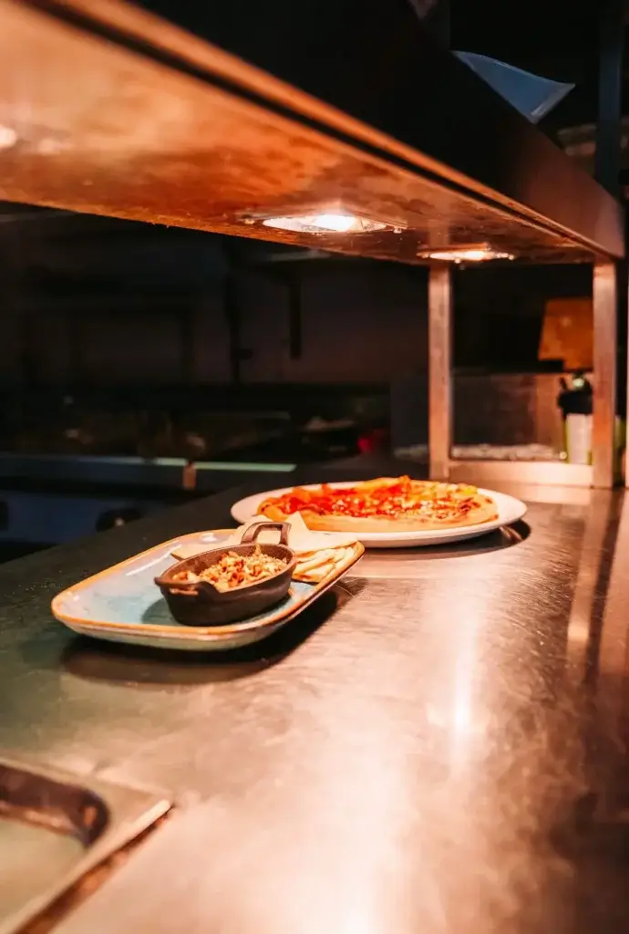 Chef’s pass with a freshly prepared pizza and small side dish under warm kitchen lights.